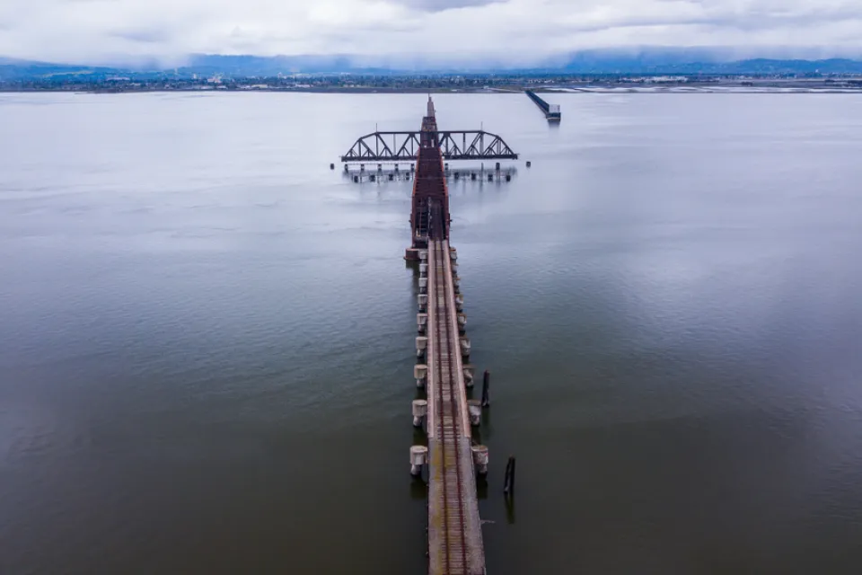 A long rail bridge, rusted and old, crosses a long stretch of water with hills and clouds on the other side.